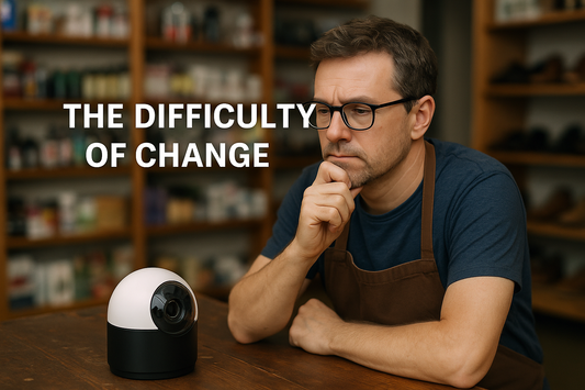 A middle-aged store owner wearing glasses and an apron sits at a counter, thoughtfully looking at a small security camera in front of him, symbolizing the challenge of outdated surveillance technology.