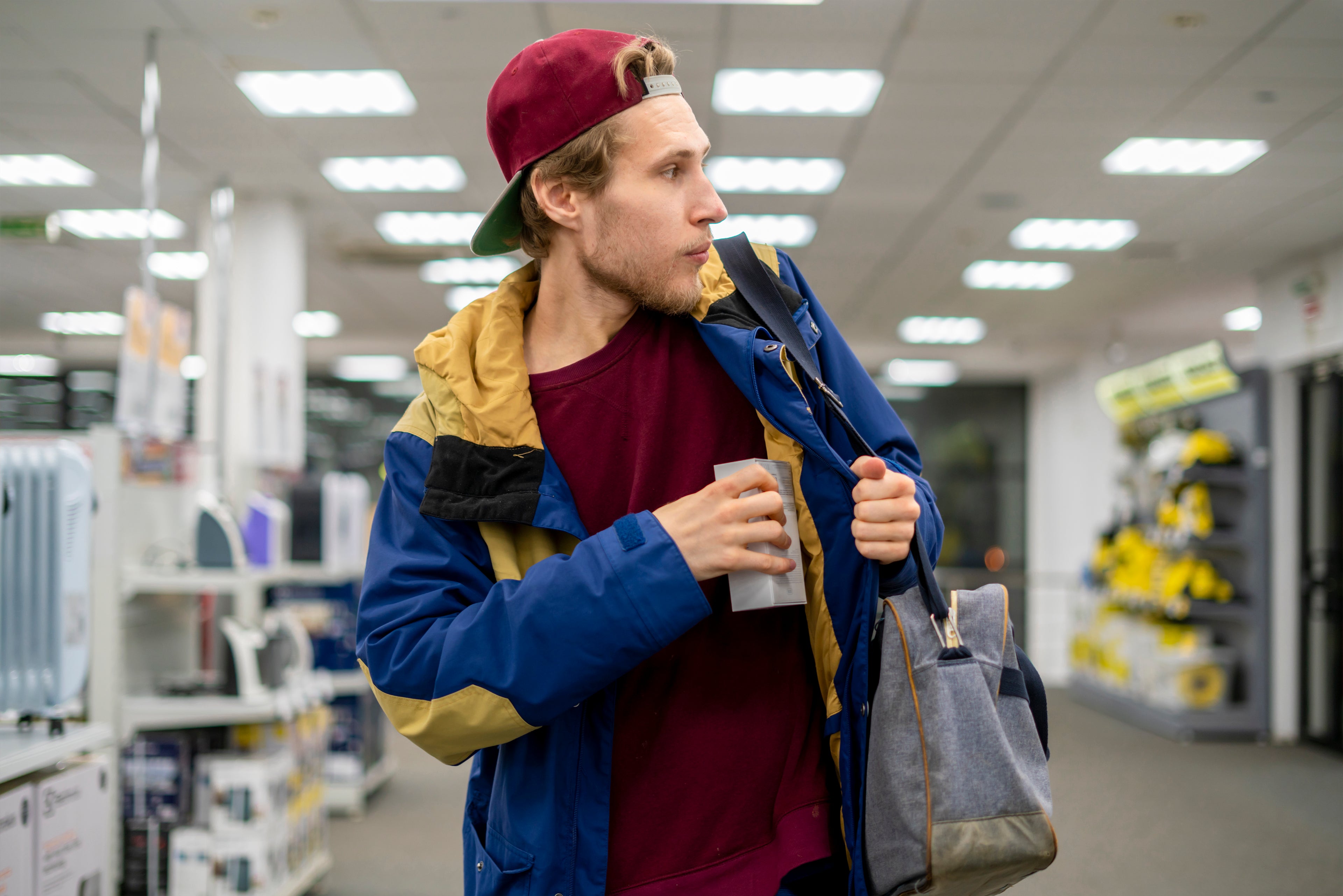 Man in a store holding a bag and a cup, wearing a blue jacket and red cap.